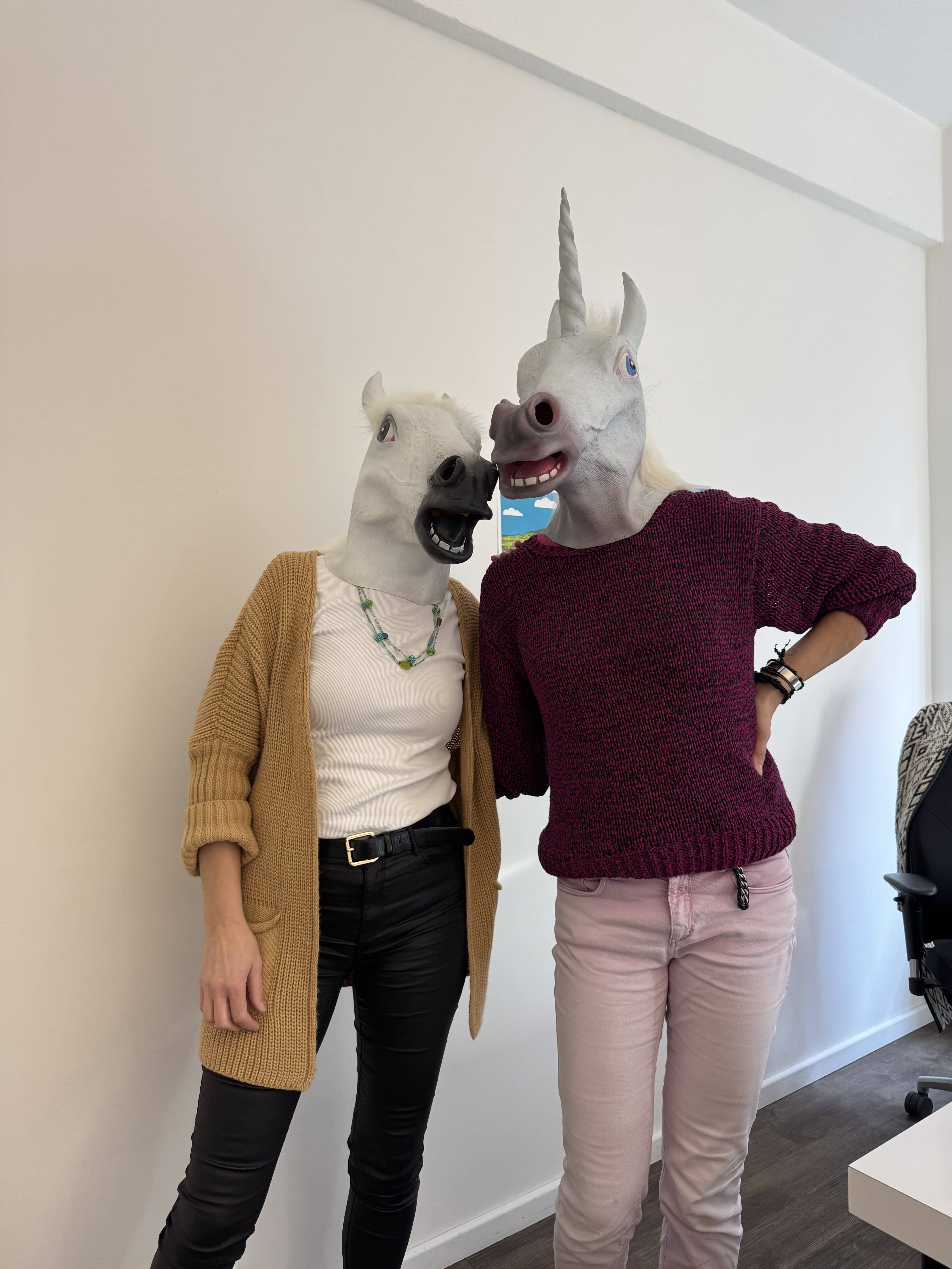 Onboarding in der Werbeagentur an Rosenmontag – kreativer Start: Zwei Personen stehen in einem Büro mit weißen Wänden und einem Schreibtisch im Hintergrund. Sie tragen Einhornmasken mit realistischen Gesichtszügen und offenen Mäulern. Die Person links trägt eine schwarze Hose, schwarze Stiefel, ein weißes T-Shirt, eine senfgelbe Strickjacke und eine grüne Halskette. Die Person rechts trägt eine rosa Hose, schwarze Stiefel, einen weinroten Strickpullover und hat eine Hand auf der Hüfte. Beide stehen entspannt nebeneinander und wirken fröhlich